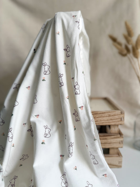 A cotton jersey fabric on an off-white background with s cute bunny rabbit and floral print in purple and pink. 