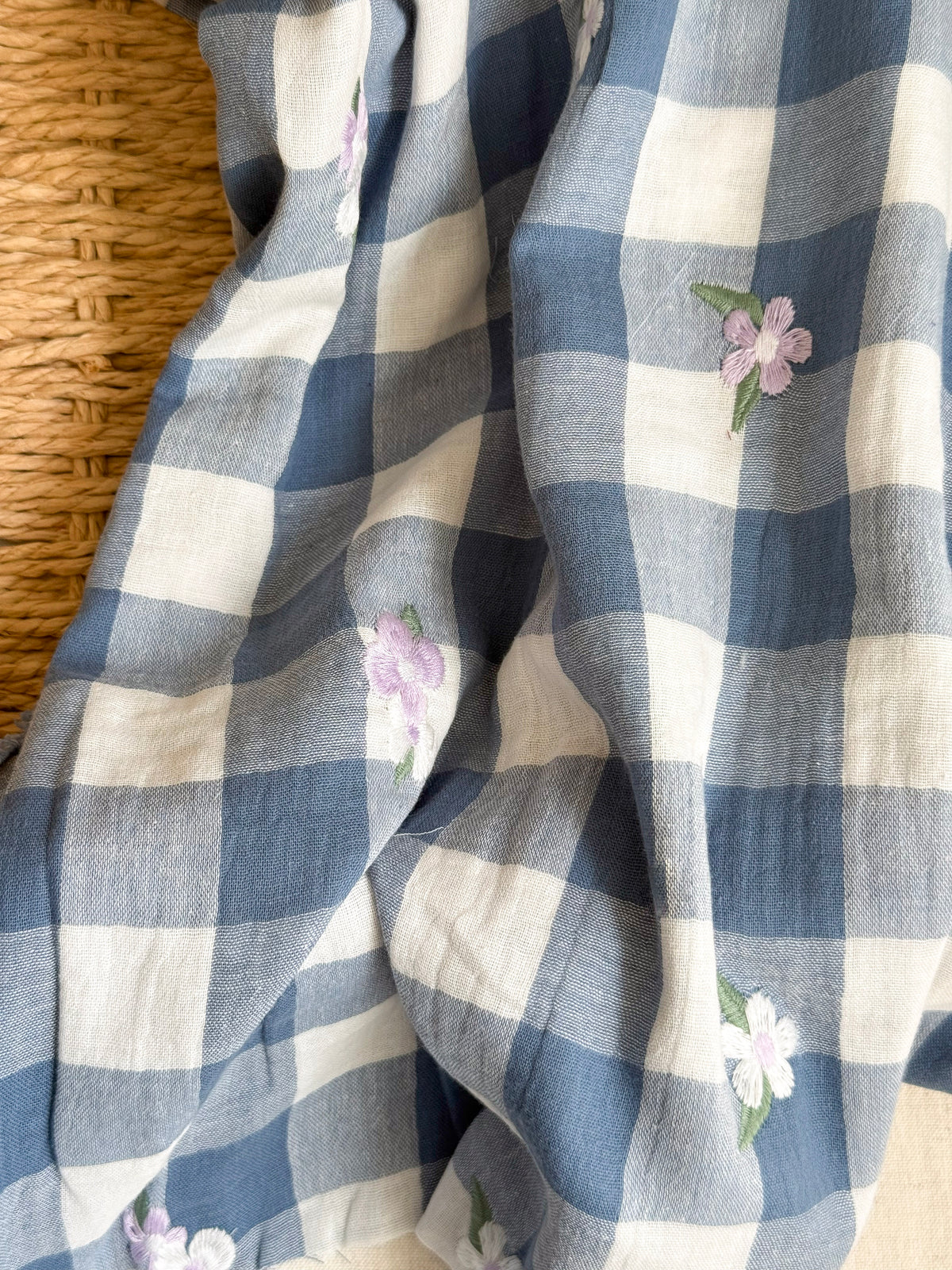 Blue and white gingham check fabric with embroidered light purple and white flowers, draped over a woven straw surface.