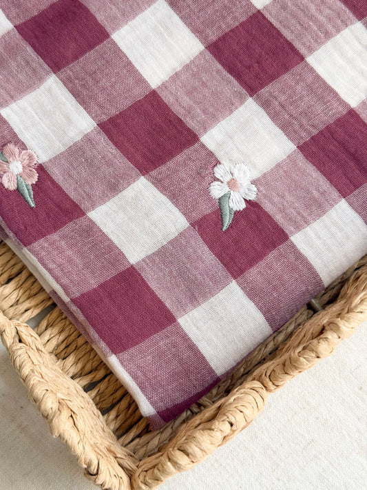 A folded checkered textile in dusty burgundy and off-white with small embroidered flowers, displayed on a woven straw tray.