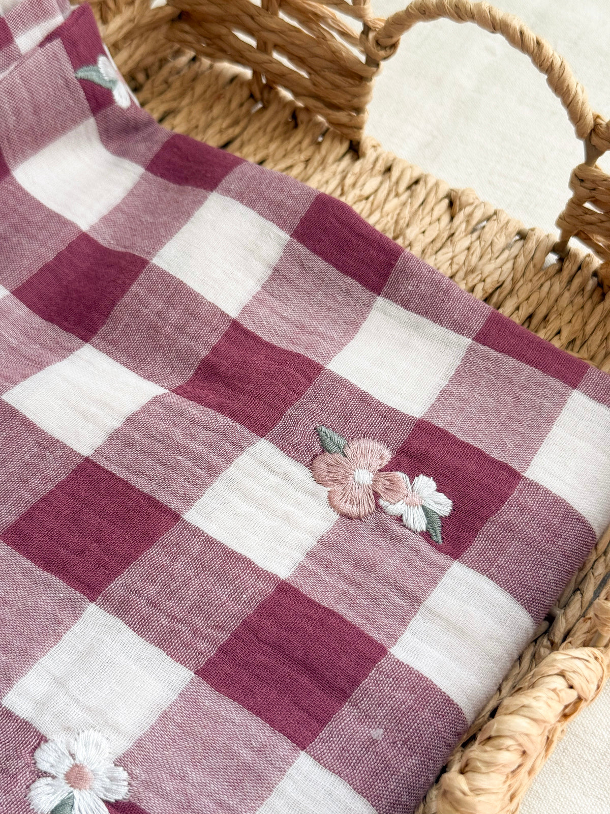 A close-up of a checkered fabric with dusty rose and white squares, decorated with small embroidered flowers, draped over a woven wicker basket.