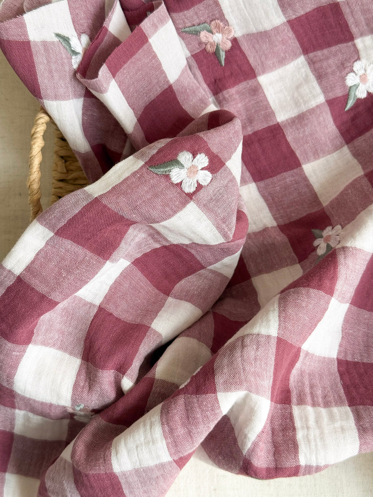 A close-up of a draped muslin fabric with a dusty rose and white gingham check pattern, decorated with small embroidered white flowers with green leaves. A portion of a woven wicker basket is visible on the left side.